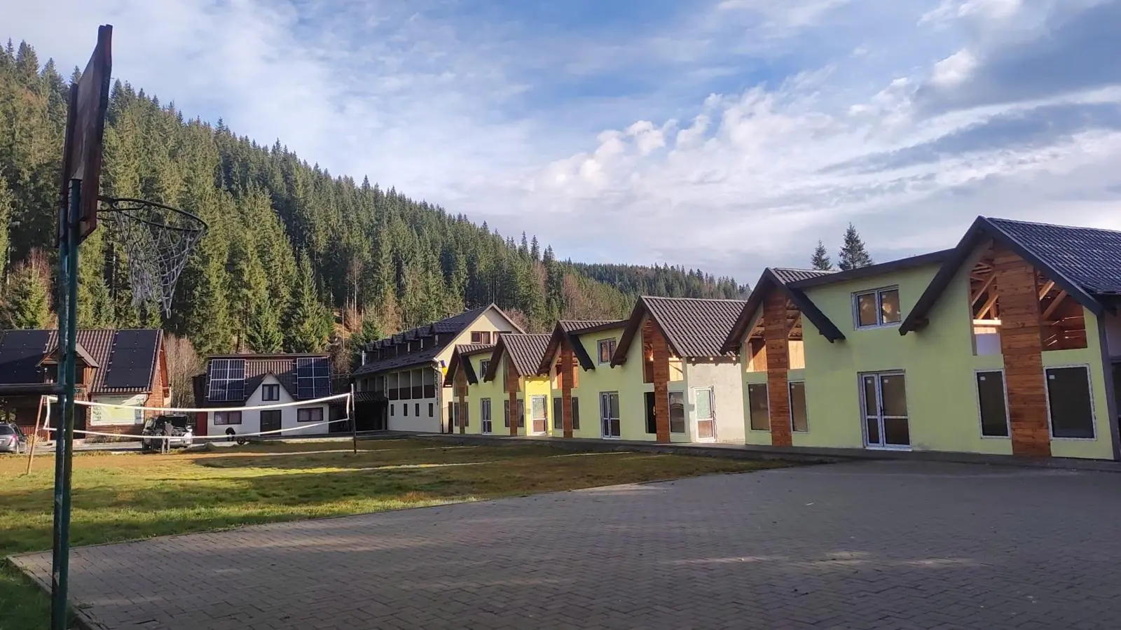 Șir de căsuțe moderne galbene, cu frontoane din lemn, lângă o curte pavată și pădurea de brazi